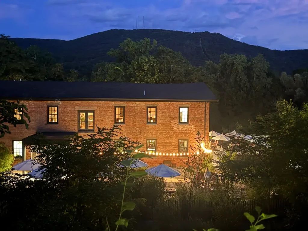 A red brick building at night with lights and sun umbrellas in a garden. Mount Beacon sits in the background against an inky blue sky.