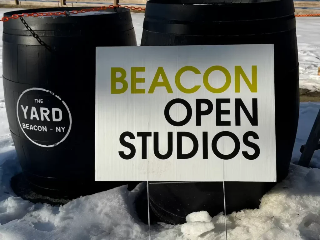 lawn sign with Beacon Open Studios in text in snow next to two black barrels.