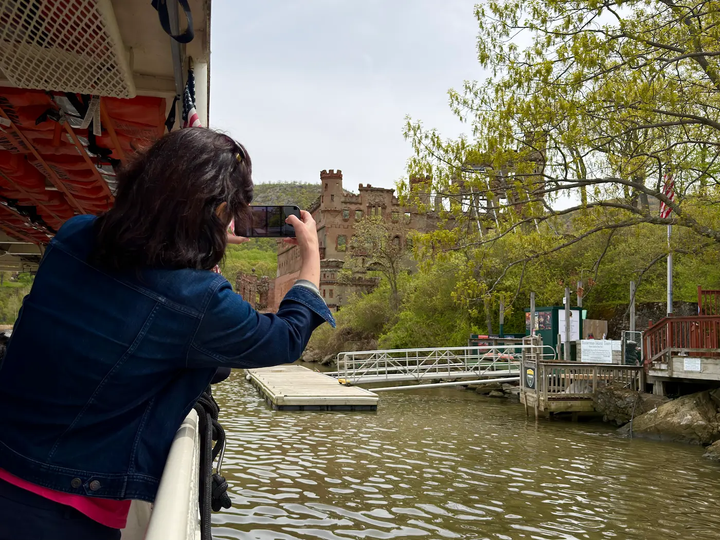 Woman leans over a boat to take a photo with her iphone of Bannerman Castle on an island.