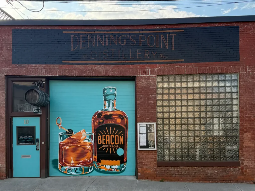 A red brick garage style building with a blue door. On it is a painted bottle of bourbon and dark colored cocktail in a glass.