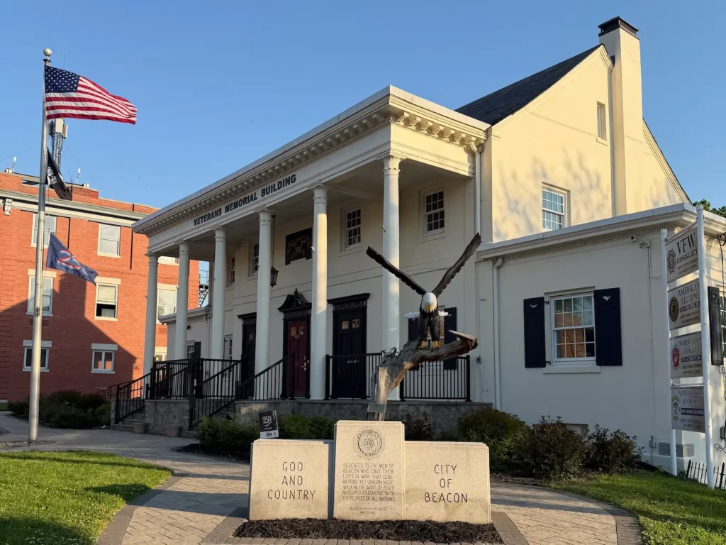 A cream colored veterans memorial building in Beacon, NY with the American flag flying high and a sculpture of an eagle flying placed to the side and front of the building. Beneath it are the words God and Country and City of Beacon.
