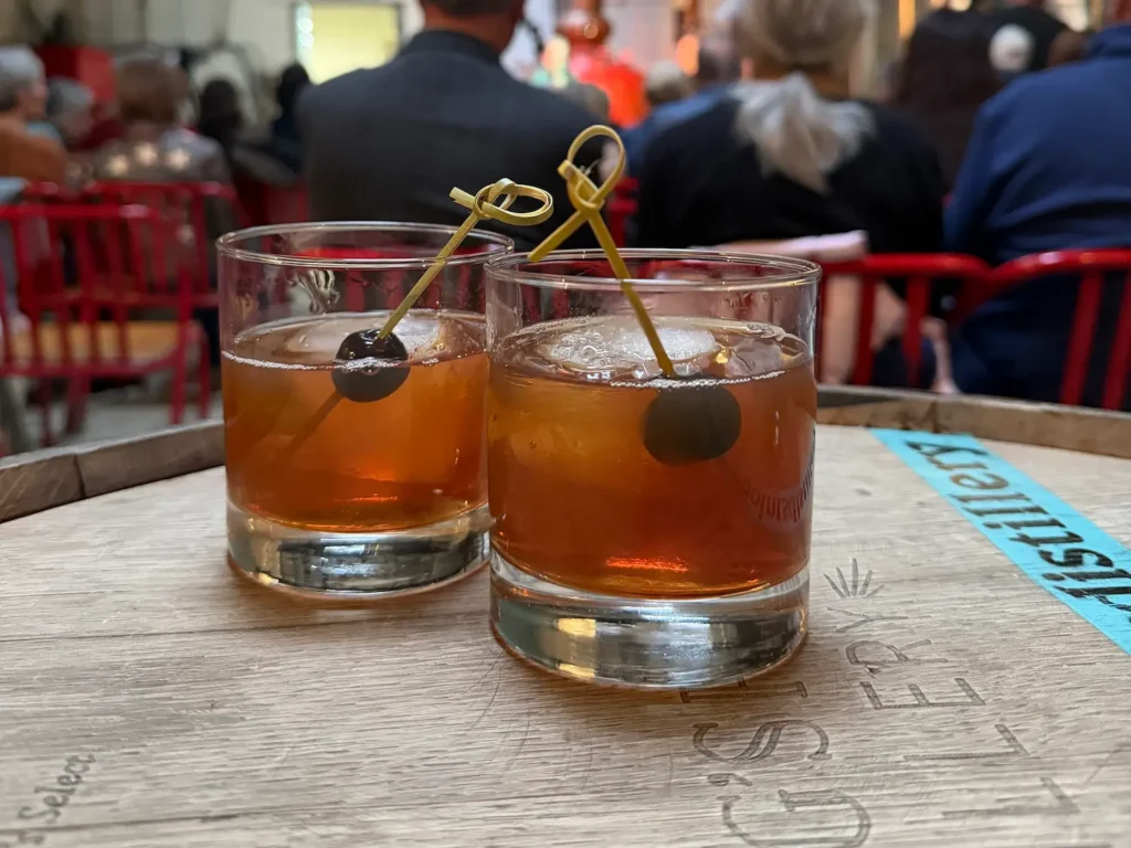 Two dark colored cocktail drinks each with a cherry sit on a barrel, with people sitting in the background.