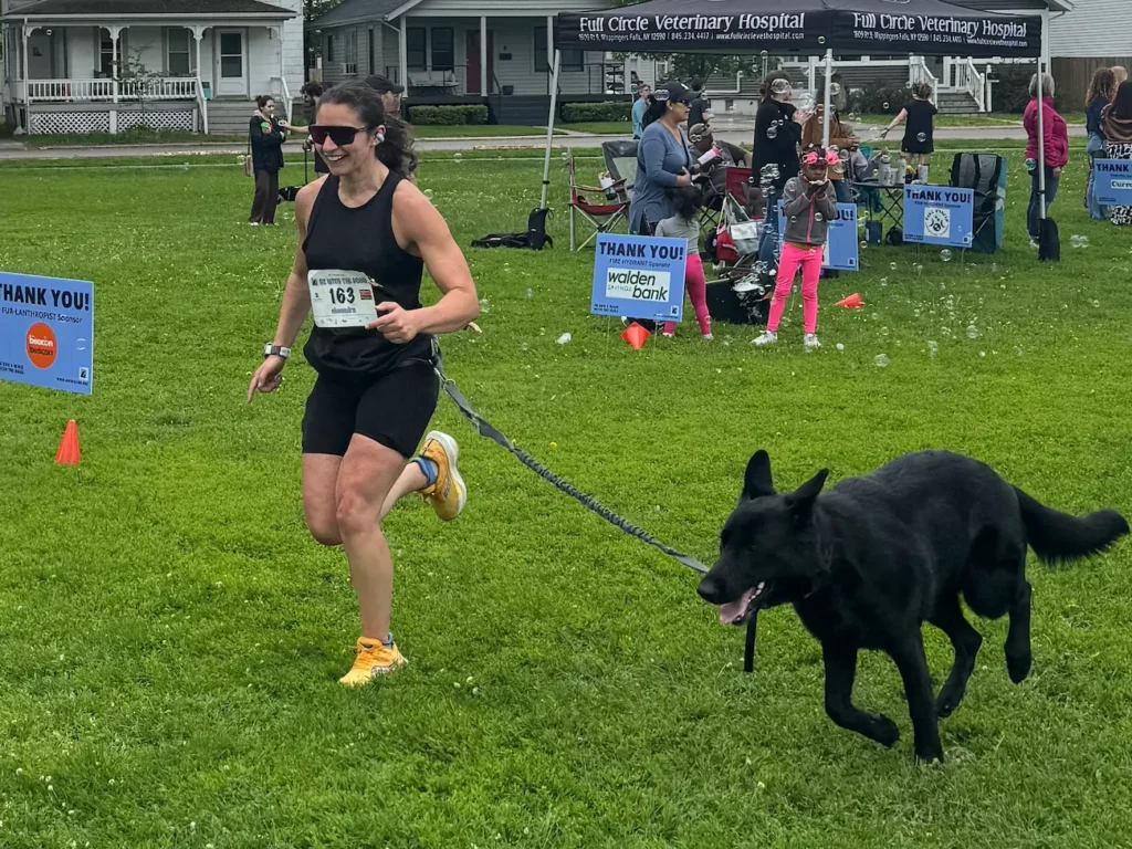 A woman wearing a black top and black shorts crosses the finishing line of a 5K race with her black dog on a leash.