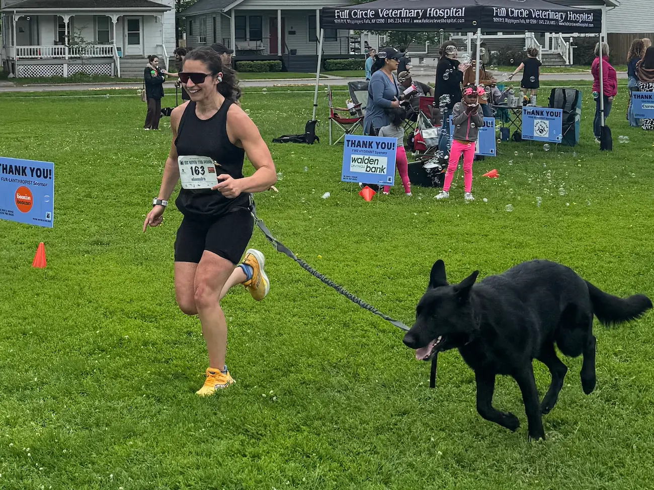 A woman wearing a black top and black shorts crosses the finishing line of a 5K race with her black dog on a leash.