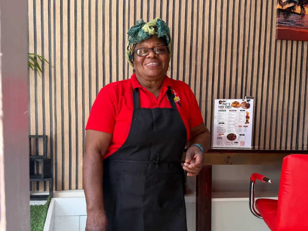 Pat of Pat's Yard Vibes in Beacon, a new Jamaican restaurant, wearing a red shirt with a black apron and a colorful headscarf.