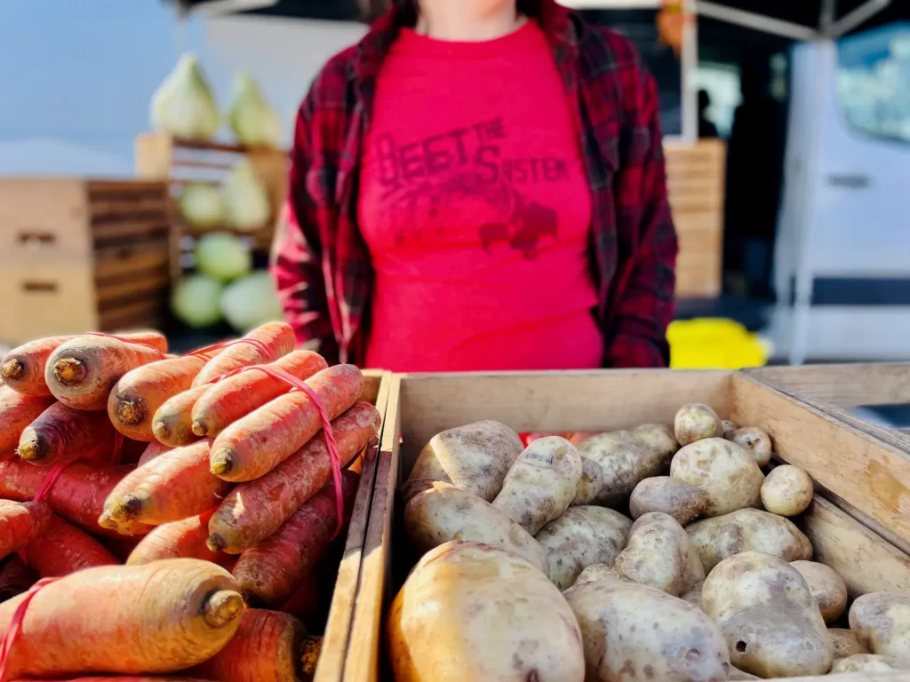 carrots and potatoes are displayed at a farmers market with a person in a red t-shirt and red plaid shirt in the background.