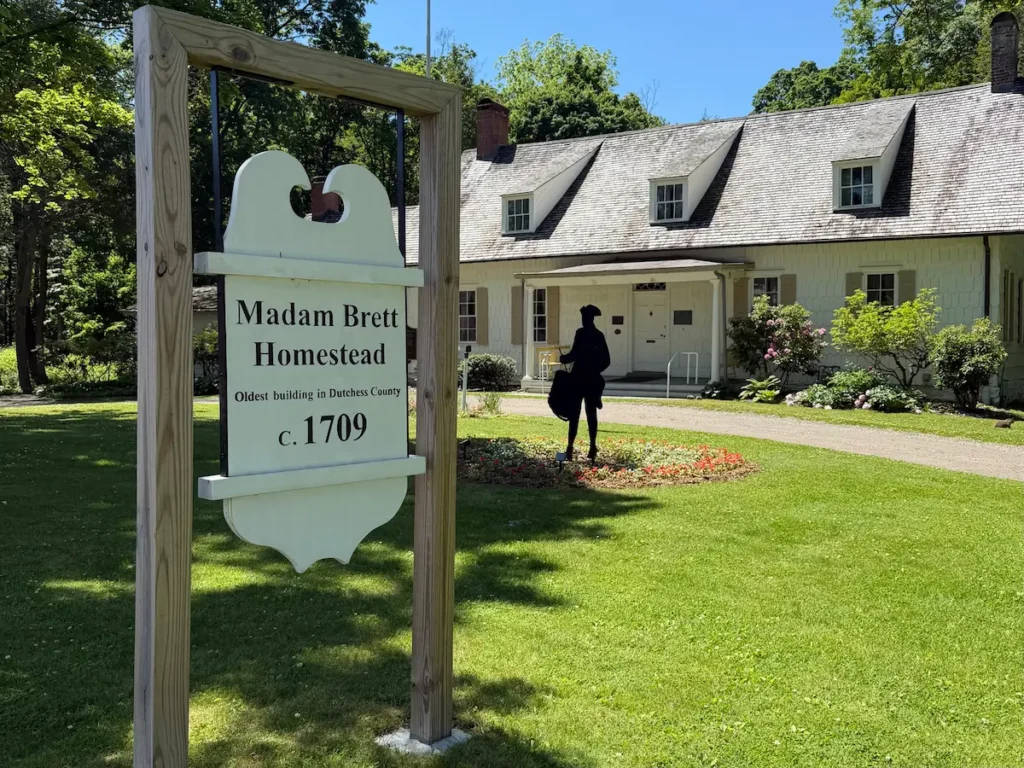 A sign in the foreground reads Madam Brett Homestead, 1709. In the background is a historic house and gardens.