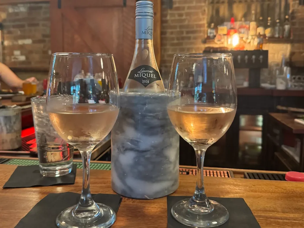 Two glasses of wine sit on a bar with a bottle of wine in between them in a marble cooler.