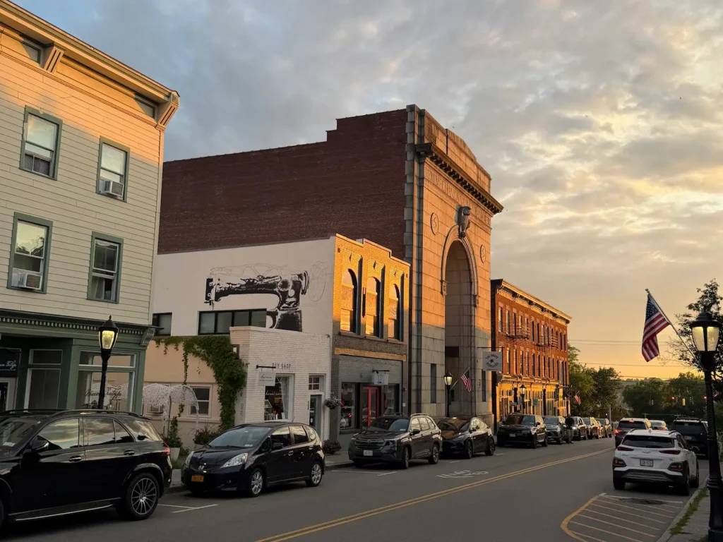Beacon NY's Main Street with Victorian buildings lining both sides of the street and cars curbside. Sunset is taking place in the background.